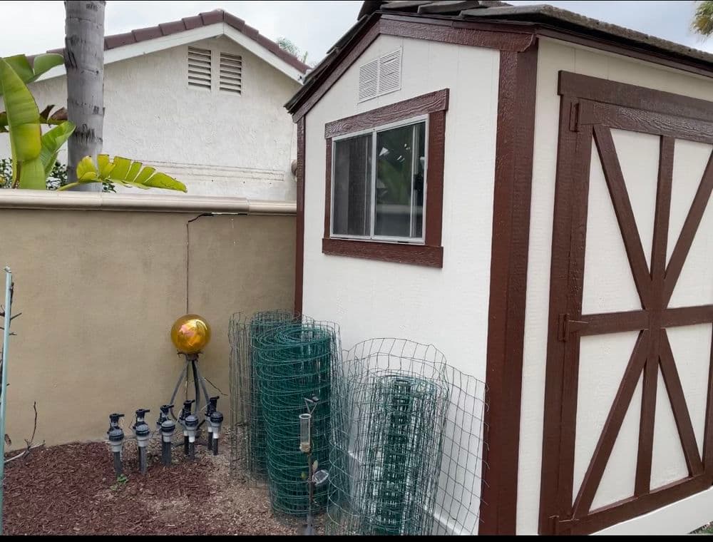 Garden shed with brown trim near fencing and landscaping, surrounded by greenery.