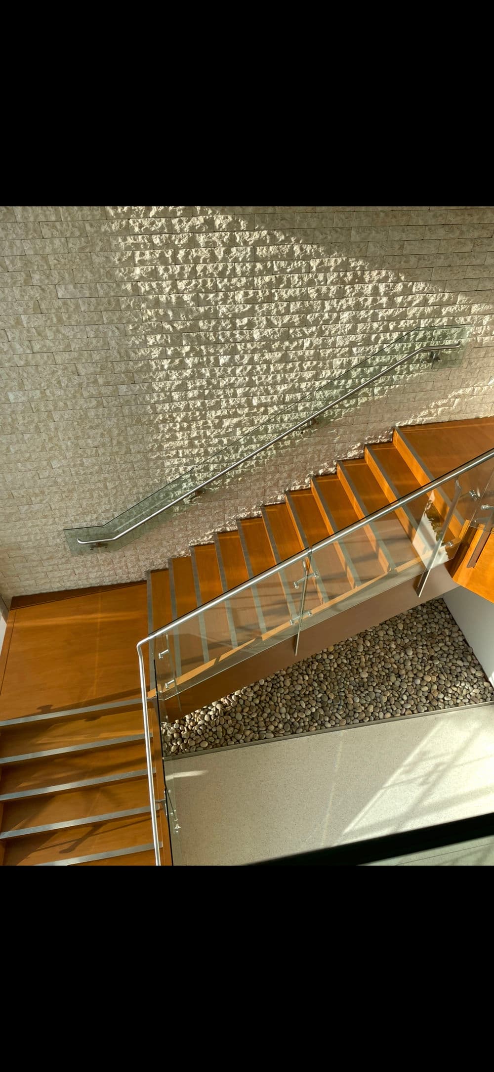 Modern wooden staircase with glass railing and textured stone wall, illuminated by sunlight.